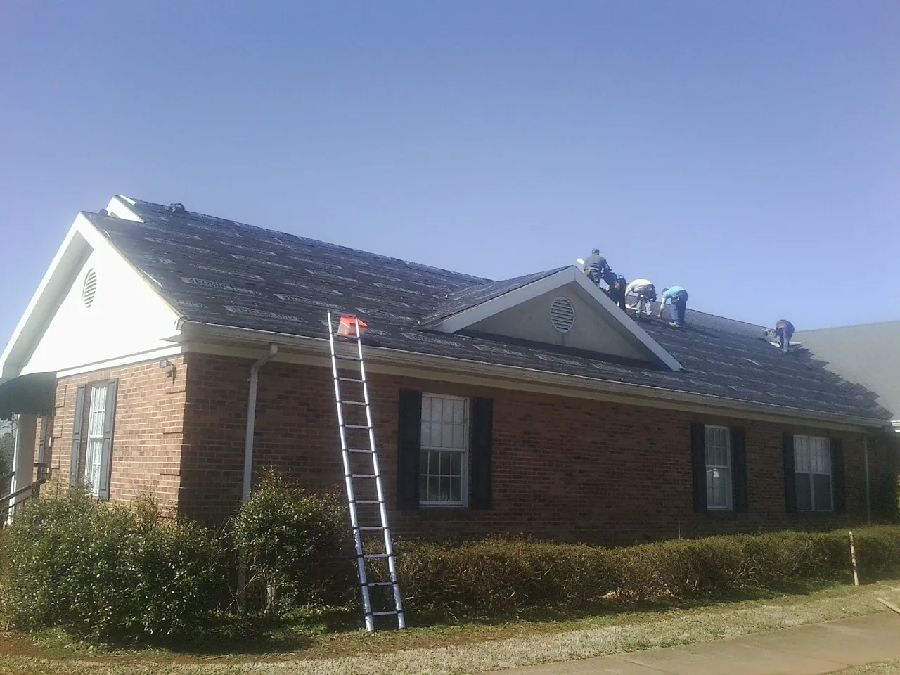 Crew tearing shingles off a brick ranch roof with a ladder set — Emergency Roof Repair work by our crew in Docena