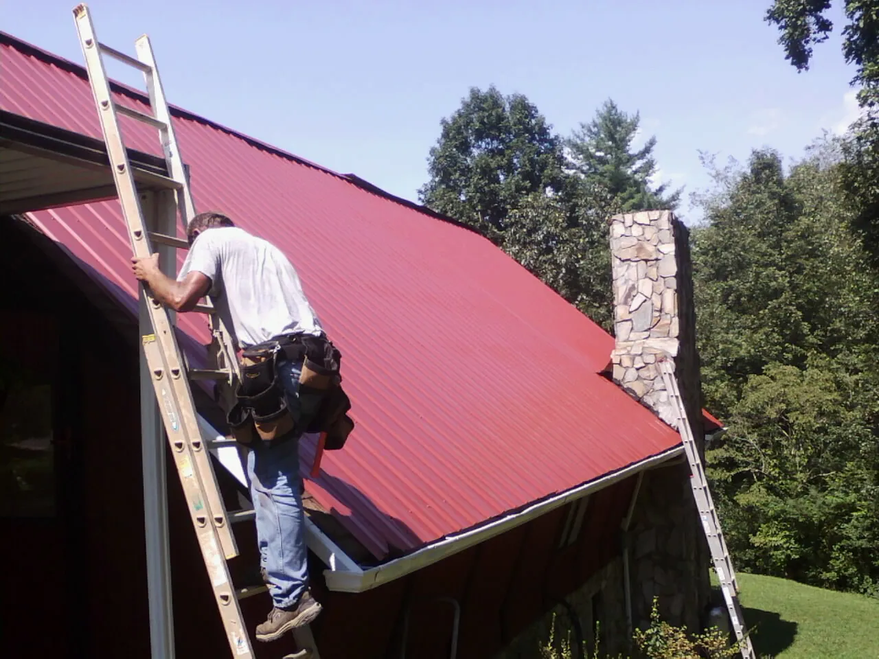 Roofer on a ladder beside a red metal roof and stone chimney — Soffit & Fascia Repair by Roofing Brothers in Docena